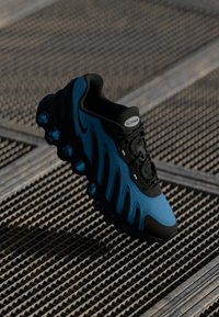 Melnas un zilas sporta apavi ar teksturētu virsmu, viļņveida dizaina elementiem un caurspīdīgu burbuļveida amortizāciju zoles daļā.