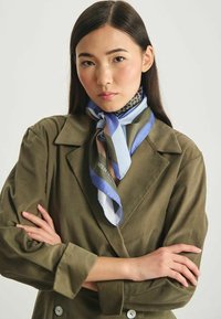 Foulard en soie présentant des motifs géométriques bleus, gris et blancs, porté autour du cou, associé à une veste verte olive.