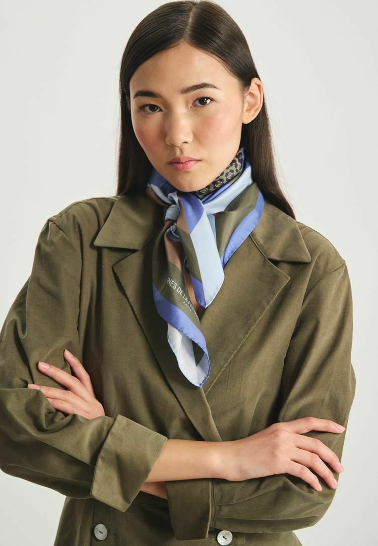 Foulard en soie présentant des motifs géométriques bleus, gris et blancs, porté autour du cou, associé à une veste verte olive.