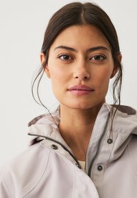 Chaqueta beige ligera con capucha, que presenta un cierre con cremallera en la parte delantera, herrajes plateados, una marca sutil y una tela suave y texturizada.
