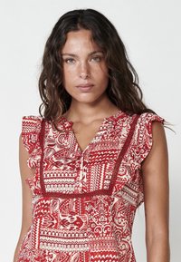 Blusa a righe rosse e bianche con scollo arricciato, maniche corte e chiusura con bottoni. Presenta disegni intricati e rifiniture a contrasto.