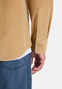 Πουκάμισο από κορντουρί σε ανοιχτό καφέ χρώμα με κάθετες ραβδώσεις, μανσέτες με κουμπιά και καμπυλωτό τελείωμα. Φορεμένο πάνω από λευκό T-shirt και συνδυασμένο με μπλε παντελόνι.