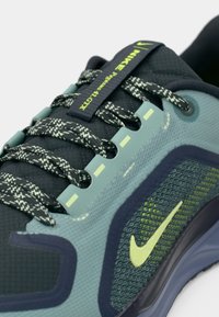 Sneaker z ciemnozieloną i jasnoniebieską siateczką, z teksturowanymi akcentami, odblaskowymi sznurówkami oraz żółtym logo Nike na boku.