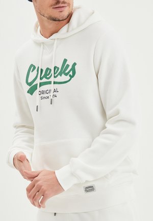 Sudadera blanca con un estampado gráfico verde que dice "Creeks Original Desde 1974". Puños acanalados, bolsillo tipo canguro y etiqueta de logo en el dobladillo.