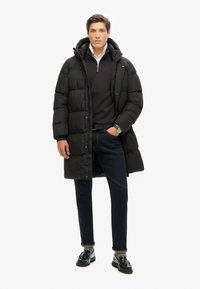 Pardessus gonflé nero con cappuccio, chiusura a zip e cuciture in vista. Indossato sopra un maglione scuro e abbinato a jeans scuri e scarpe nere.