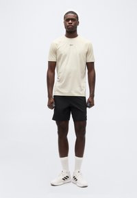 Νέος άνδρας που στέκεται, κρατώντας μικρά αλτήρες, φοράει μπεζ μπλουζάκι BOSS, μαύρες σορτς, λευκές κάλτσες και αθλητικά παπούτσια Adidas σε απλό φόντο.