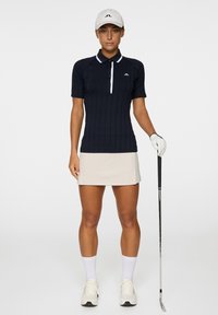 Polo shirt a coste navy con accenti bianchi, maniche corte, bottoni frontali, abbinato a una gonna beige. La modella tiene una mazza da golf e indossa un cappellino.