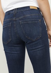Temno modre kavbojke iz denima s srednjim pasom, ki imajo dva zadnja žepa, neopazno ob stitching in usnjeno oznako blagovne znamke na pasu.