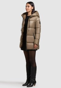 Dikke, lichtbruine parka met een capuchon, ritssluiting aan de voorkant, twee zijzakken en zwarte details. Draagt over een zwart jurkje met kniehoge zwarte laarzen.