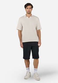 Beige ribbet kortærmet polo skjorte med V-hals, parret med sorte knælange shorts og hvide sneakers med brand detaljer.