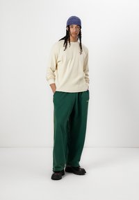 Cream long-sleeve shirt, embroidered detail, paired with green sweatpants featuring side stripes. Worn with a blue ribbed beanie and black shoes.