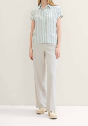 Kobieta ubrana w jasnoniebieską i białą koszulę z krótkim rękawem w pionowe paski, jasnobeżowe szerokie spodnie oraz beżowe buty.