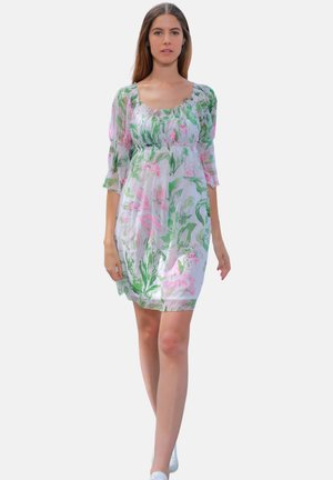 Femme marchant vers l'avant portant une robe blanche longueur genou avec des motifs floraux verts et roses et des manches trois-quarts transparentes.