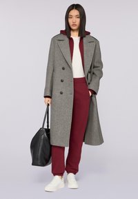 Grauer Wollmantel mit doppelreihigen Knöpfen, getragen über einem burgunderfarbenen Hoodie und passenden Hosen. Weiße Sneaker und eine schwarze Tasche vervollständigen den Look.