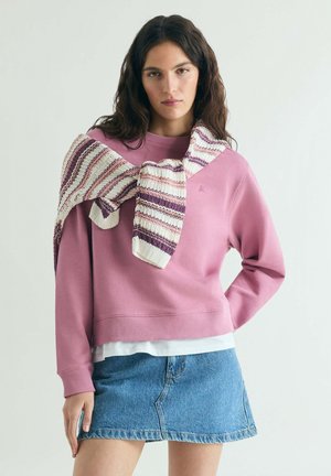Mujer con sudadera rosa, mini falda de mezclilla azul, camiseta blanca debajo y un suéter de punto a rayas sobre los hombros.