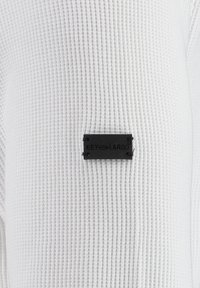 Tissu blanc texturé avec un motif en nid d'abeille. Comprend une petite étiquette rectangulaire noire avec "KEY LARGO" embossé. Les coutures sont visibles.
