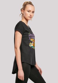 Black short-sleeve t-shirt featuring colorful cartoon graphic with "The Amazing Scooby" text. Relaxed fit with a slightly longer back.