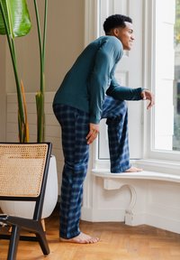 Blauwe lange mouwen shirt gecombineerd met geruite pyjamabroek, met groene en blauwe tinten. De persoon staat op een vensterbank.