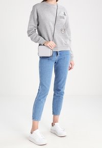 Grijze sweatshirt met patches, blauwe spijkerbroek en witte sneakers. Een kleine witte schoudertas met een zwarte band maakt de outfit compleet.