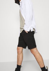 Bílá košile na knoflíky přes zelené a bílé pruhované tričko, v kombinaci s černými šortkami a tmavými ponožkami, předvádějící volnočasový outfit.