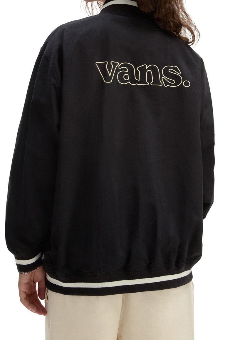 Osoba ubrana w czarną kurtkę Vans z białymi paskami na mankietach i dolnym brzegu, w beżowych spodniach, pokazana z tyłu z widocznym logo "vans.".
