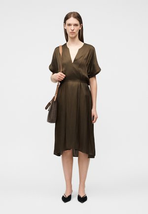 Γυναίκα που φοράει midi φόρεμα σε σκούρο ελιά με V-λαιμόκοψη και κοντά μανίκια, μαύρα μυτερά παπούτσια και κρατάει καφέ τσάντα ώμου.