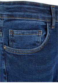 Modre kavbojke z zadnjim žepom, ki imajo oranžne šive in srebrn gumb. Tkanina se zdi teksturirana in trpežna.