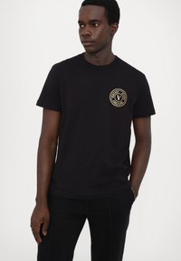 Tricou din bumbac negru, cu guler rotund și mâneci scurte. Partea din față prezintă un imprimat cu logo circular aurit. Design slim fit.