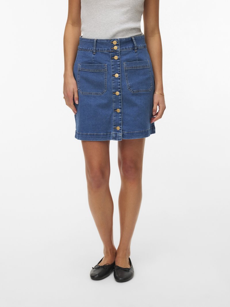 Femme portant une jupe boutonnée en denim bleu avec deux poches avant, associée à des ballerines noires et un haut léger sans manches.