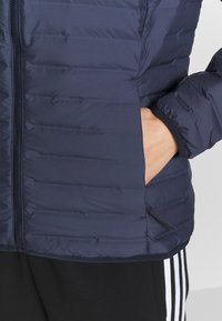 Main dans la poche d'une veste matelassée bleu marine avec fermeture éclair, associée à un pantalon noir avec des bandes blanches sur les côtés.