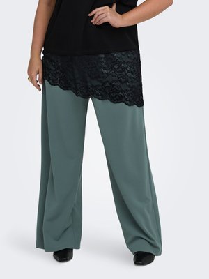 Femme portant un pantalon vert à jambes larges associé à un haut noir avec un ourlet en dentelle noire, debout avec une main sur la hanche.