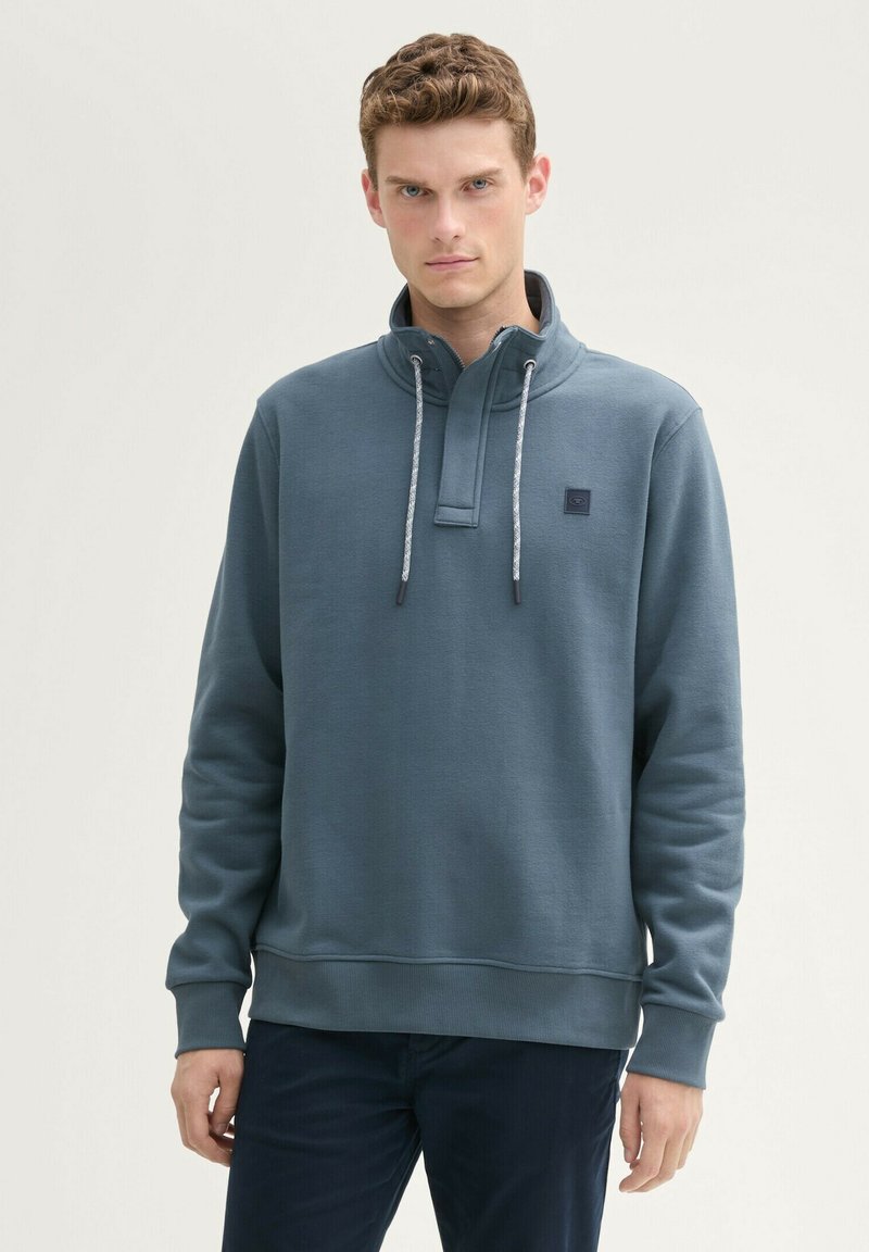 Sweatshirt gris avec un col haut, fermeture éclair mi-hauteur et accents avec cordon. Fabriqué en tissu doux, avec un petit écusson logo sur la poitrine.