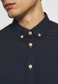 Navy blue button-up shirt featuring a pointed collar, wooden buttons, and a textured fabric with visible stitching details along the placket.
