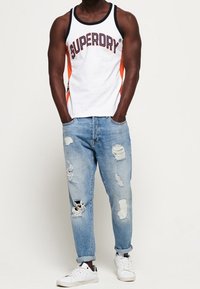 Camiseta sin mangas blanca con "SUPERDRY" en azul marino y naranja, combinada con vaqueros desgarrados azul claro y zapatillas blancas. Atuendo casual y deportivo.