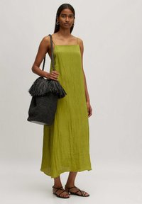 Grünes Leinen-Midi-Kleid mit Spaghetti-Trägern, strukturiertem Stoff, kombiniert mit einer gewebten schwarzen Bucket-Bag mit Fransenakzent. Schwarze flache Sandalen.