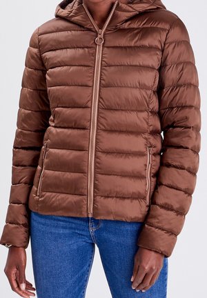 Gros plan d'une personne portant une veste matelassée marron zippée avec capuche et un jean bleu, les mains détendues sur les côtés.