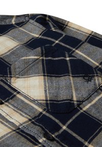 Ravvicinato di tessuto a quadri blu navy e beige con una piccola tasca cucita e dettagli di cucitura visibili.