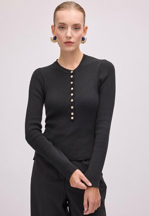 Femme portant un haut noir cintré à manches longues avec des boutons crème sur le devant et des boucles d’oreilles bleues et dorées imposantes sur fond uni.