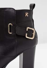 Sort læder ankelstøvle med blokhæl, guldfarvet spænde og lille guld bogstav "R" detalje på siden.