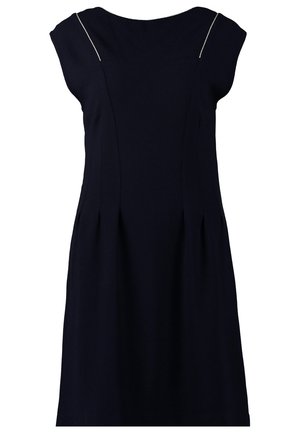 Femme en robe noire sans manches avec un détail de fermeture éclair à l'avant, collants noirs, bottines noires, tenant une petite pochette argentée.