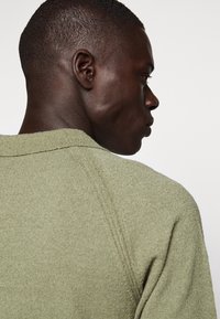 Zelený pletený svetr s texturovanou tkaninou a vroubkovaným límcem, zobrazený z zadní strany, zdůrazňující tvar a detaily švů.