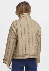 Quiltet beige jakke med høj krave, synlige syninger og en boxy silhuet. Har fremhævede sømme og et struktureret bagdesign.