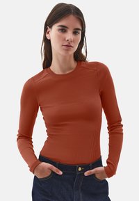 Joven mujer con cabello largo y marrón, vistiendo una camiseta ajustada de manga larga de color óxido y jeans de mezclilla oscuros, con las manos en los bolsillos.