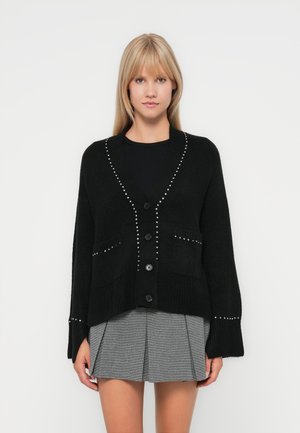 Svart strikket cardigan med V-hals, fem knapper og sølvfargede nagledekorasjoner. Har to frontlommer og ribbestrikkede mansjetter.