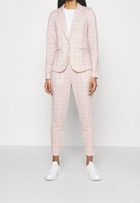 Ljusrosa kostym med orange prickar, bestående av en välsittande kavaj och avsmalnande byxor. Matchad med vita sneakers. Slät tygtextur.