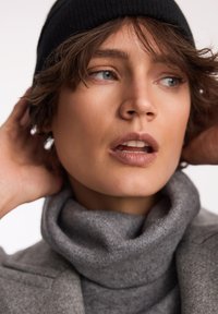 Szary sweter typu golf o miękkiej fakturze i wysokim kołnierzu, zestawiony z czapką w prążki z czarnej dzianiny. Skupienie na twarzy i szczegółach fryzury.