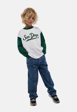 Chemise à manches longues blanche et verte avec l'impression "San Diego", portée avec un jean bleu ample. L'enfant se tient avec les mains dans les poches, portant des baskets.