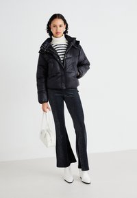 Schwarze Daunenjacke mit abnehmbarem Kapuzenaufsatz, gestepptem Muster, kombiniert mit einem gestreiften Pullover, glitzernden Schlaghosen sowie einer weißen Handtasche und Stiefeln.