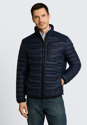 Homme portant une veste matelassée bleu marine, debout avec une main dans la poche devant un fond clair uni.