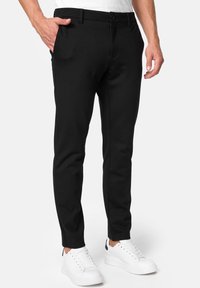 Pantalon noir ajusté en tissu lisse, avec poches avant et fermeture par bouton. Porté avec des baskets blanches pour un contraste.